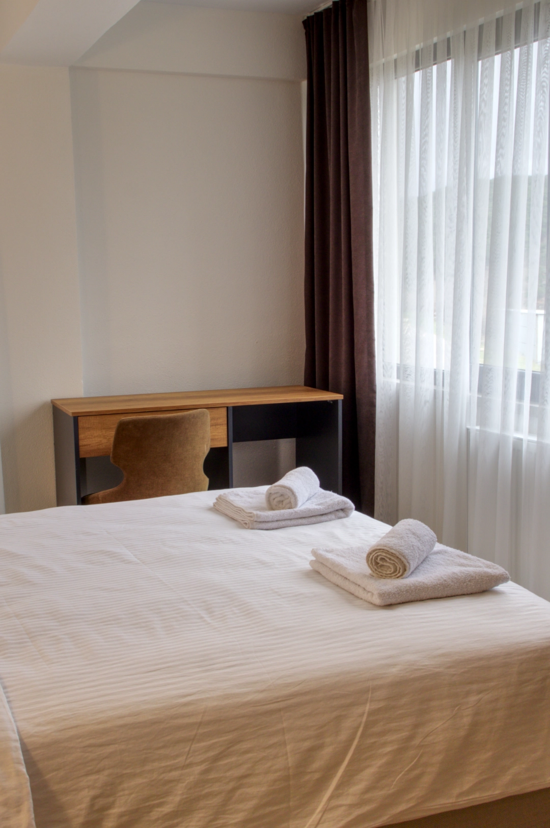 Beyaz nevresimli bir yatak, ahşap çalışma masası ve geniş pencerelere sahip aydınlık bir otel odası.